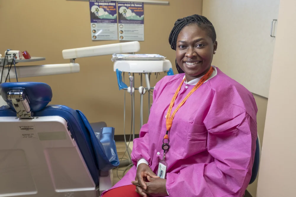 Smiling dental professional in pink gown seated beside exam chair